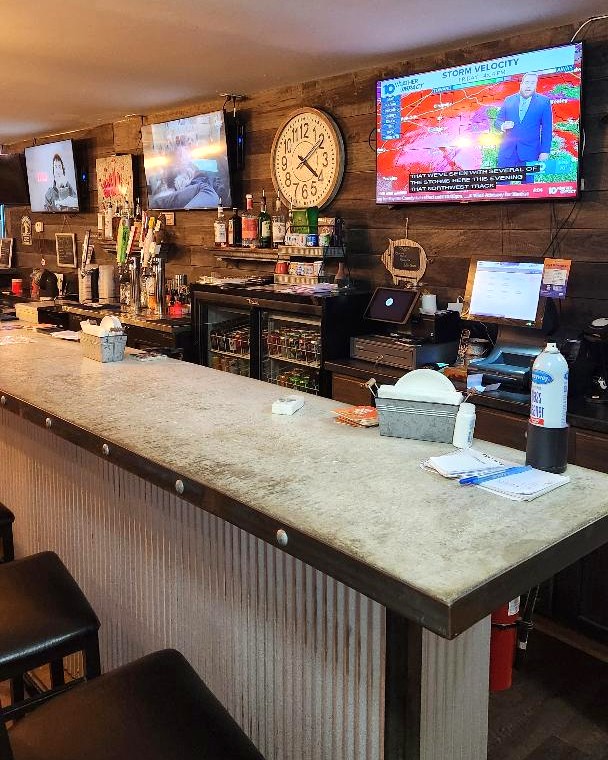 The bar service area with a register screen, glass-door cooler, taps and wall-mounted televisions
